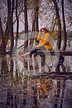 A young girl in a yellow sweater sits on the bridge over the lake and catches fish