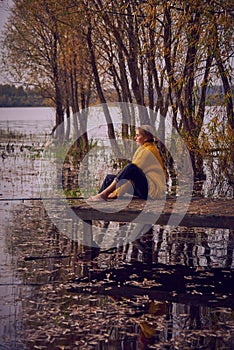 A young girl in a yellow sweater sits on the bridge over the lake and catches fish