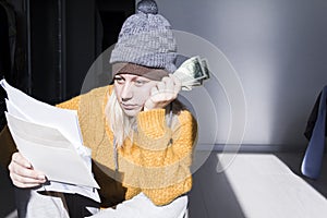 A young girl in a yellow sweater and two hats is horrified to study large bills, taxes and utility bills