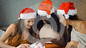 Young girls of Europe, Asia and Africa in red New Year`s clothes and hats sit on the bed at the Christmas tree, give gifts and