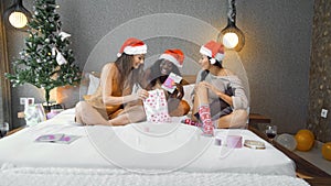 Young girls of Europe, Asia and Africa in red New Year`s clothes and hats sit on the bed at the Christmas tree, give gifts and