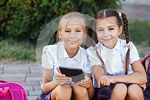 Young girls reading an ebook. Pupils of primary school. Girls with backpacks near building outdoors. Beginning of lessons. First d