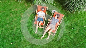 Young girls relax in summer garden in sunbed deckchairs on grass, women friends have fun outdoors in green park on weekend