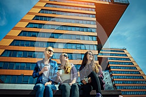 Young girls in sunglasses sitting on a city street and drink coffee to take away. Three cute women have a coffee break together