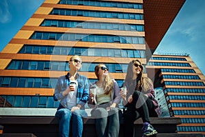 Young girls in sunglasses sitting on a city street and drink coffee to take away. Three cute women have a coffee break together