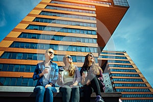 Young girls in sunglasses sitting on a city street and drink coffee to take away. Three cute women have a coffee break together