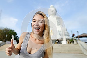 Young gladden woman showing thumbs up, white Buddha statue in Phuket in background.