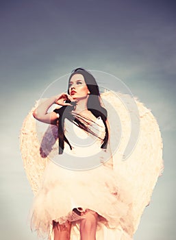 Young glamorous woman in a white dress with a large white angel wings on the background of a dramatic sunset sky.