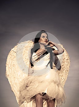 Young glamorous woman in a white dress with a large white angel wings on the background of a dramatic sunset sky.