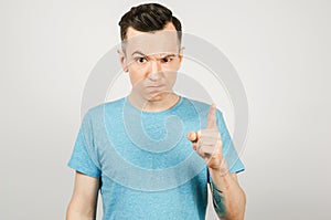 Young gloomy threaten man with a finger, dressed in a blue t-shirt on a light background. Close up portrait