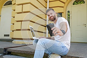 Young good looking bearded guy is sitting outdoors on stairs in front of his house working on laptop. he is smiling and drinking