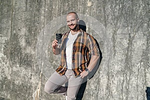 Young good looking bold bearded freelancer standing outdoors against grey modern loft wall holding coffee in his hand and