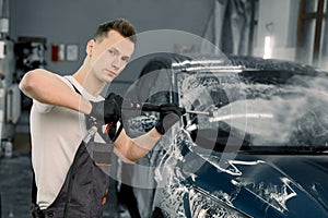 Young good-looking man worker washing luxury dark blue car on a car wash, looking at camera and holding high pressure