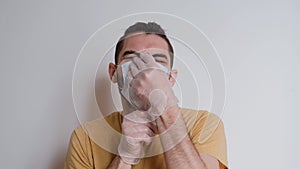 Young, good looking white Caucasian man with beard puts white protective medical mask on his face and stares intently ahead.