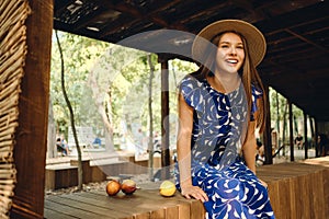 Young gorgeous smiling woman in blue dress and hat happily sitting with delicious peaches on wooden fence in park