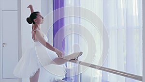 Young graceful ballerina is stretching tilting to her leg near the barre stand.