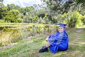Young Graduate Proudly Sitting By A Pond With Cap And Gown