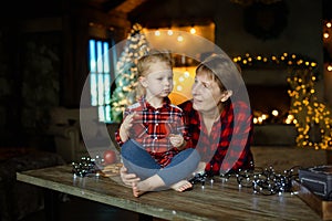 A young grandmother with her little granddaughter who sits on a table and eats sweets from a Christmas gift.