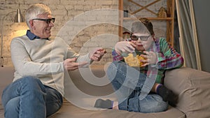 Young greedy boy takes a bowl of chips from his grandfather. Elderly man and a young fat boy are sitting on the couch in