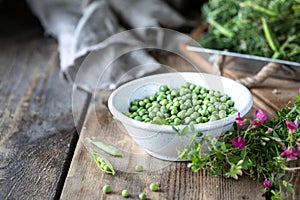 Young green peas in white bowl on wooden background. Pea flowers and daisy flowers on the table