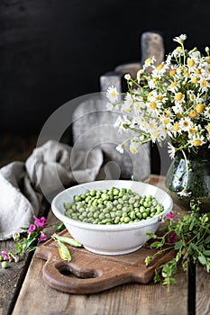 Young green peas in white bowl on wooden background. Pea flowers and daisy flowers on the table