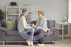 Young grown daughter and old senior mother holding hands sitting on home sofa