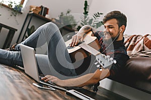 Young guitarist hipster at home sitting on the floor holding guitar earphones music browsing laptop