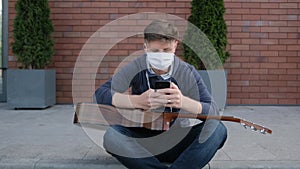 Young guitarist is sitting on the sidewalk and typing a message to his friends.