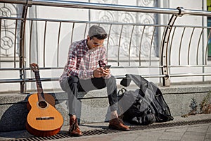 Young guitarist sitting and using phone in the street