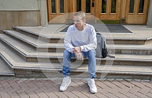 A young guy of 20-25 years old in summer clothes with a backpack is sitting on the steps at the entrance to the institution. Close