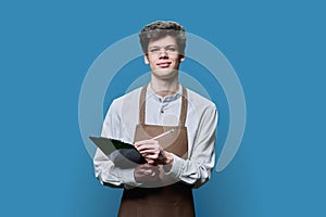 Young guy in apron making notes on clipboard, on blue studio background