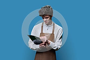 Young guy in apron making notes on clipboard, on blue studio background