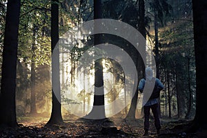 Young guy with a backpack standing in a forest in the mist at sunrise