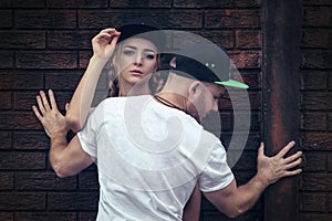 A guy in a white t-shirt shields a girl in a baseball cap against a wall