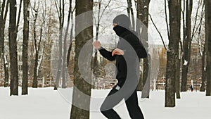 A young guy in black sports clothes and Balaclava, performing a warm-up before training in a winter snow-covered city