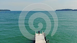 Young guy in a blue shirt and shorts stands on the pier and looks into the distance at the sea against a beautiful