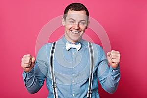 Young guy with blue shirt, suspenders and bow tie, celebrating something with his fists up, isolated on pink background. Handsome