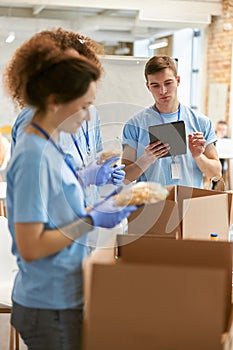 Young guy calculating using tablet pc. Volunteers in protective gloves sorting, packing food in cardboard boxes, working