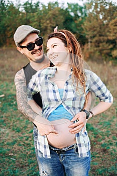 young guy in a cap solar oskah, with beard mustache, and tattooed, his pregnant girl in dreadlocks, outdoors the fall posing at t