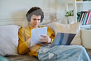 Young guy college student in headphones using laptop sitting on couch at home