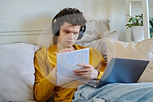 Young guy college student in headphones using laptop sitting on couch at home
