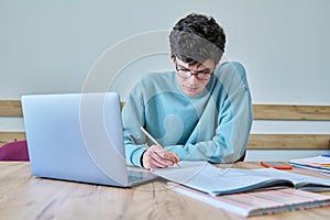 Young guy college student sitting at desk in classroom, using laptop, writing in notebook