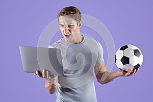 Young guy disappointed at his soccer team defeat, watching football game on laptop computer, lilac studio background
