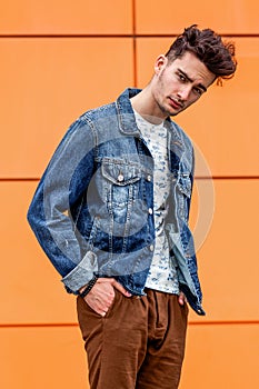 Young guy dressed in casual clothes brown trousers and jeans jacket poses in the street on the background of a orange