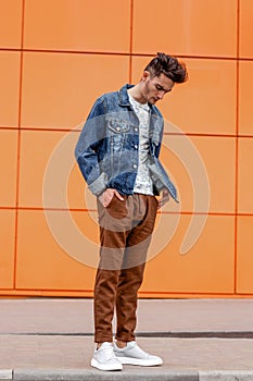Young guy dressed in casual clothes brown trousers and jeans jacket poses in the street on the background of a orange