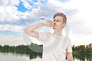 Young guy drinks fresh water against of the lake and the forest background. summer thirst. Guy wants to drink