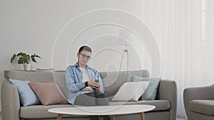 Young guy in eyeglasses watching tv at home, sitting on sofa and switching channels