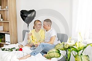 A young guy and a girl in the bedroom with glasses of wine, a balloon and flowers celebrating Valentine& x27;s Day or