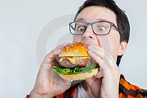 A young guy with glasses holding a fresh Burger. A very hungry student eats fast food. Hot helpful food. The concept of gluttony a