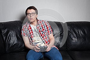 Young guy with glasses and red shirt playing video games on the joystick, sitting on a black leather sofa